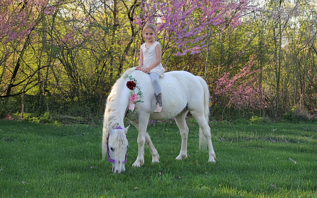 Young girl riding a white pony in a park with trees and flowers in the background