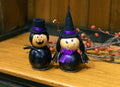 Two Halloween decorative figures shaped like a pumpkin and a gourd, both black with painted faces, hats, and purple ribbon accents, displayed on a wooden cookie.