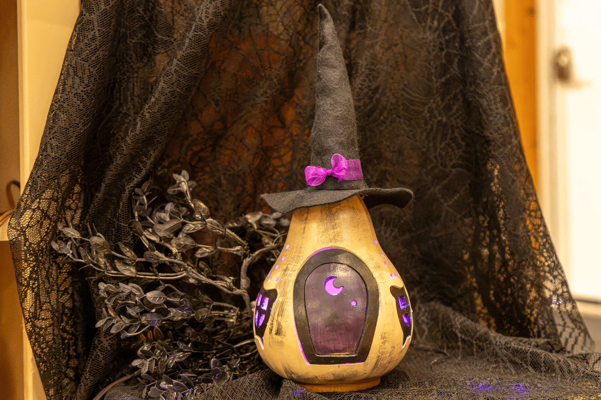 A decorative gourd lantern shaped like a witch's house, wearing a black pointed hat with a purple bow, set against a backdrop of black lace fabric and dried black foliage.