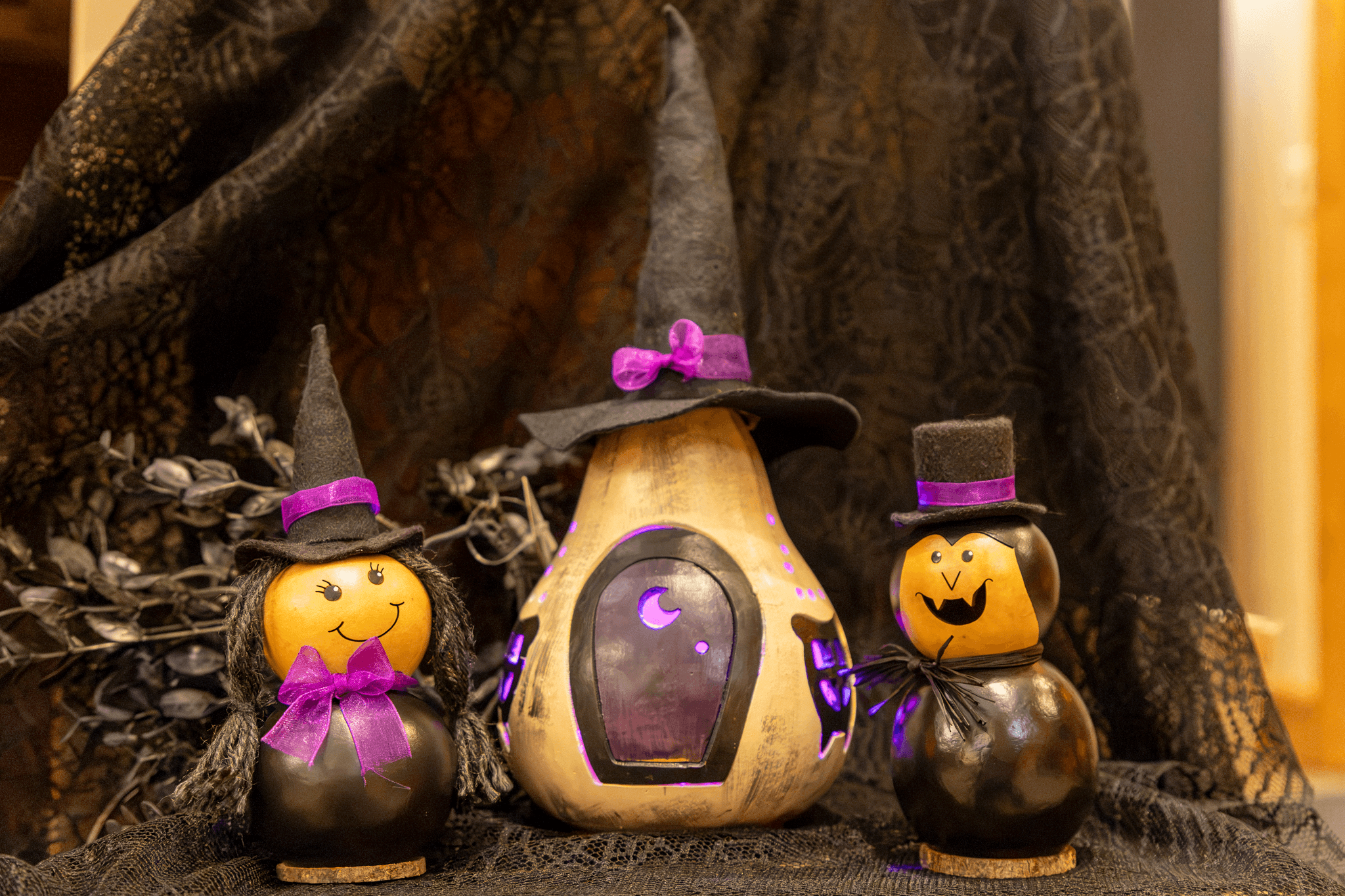 Three Halloween decorations arranged together: a central painted gourd with a witch hat and purple door, flanked by two small witch and pumpkin figures with purple bows and hats, set against dark draped fabric.