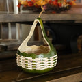 A decorative handcrafted gourd basket with a green body, natural gourd handle, and woven white reed, displayed on a dark wooden surface.