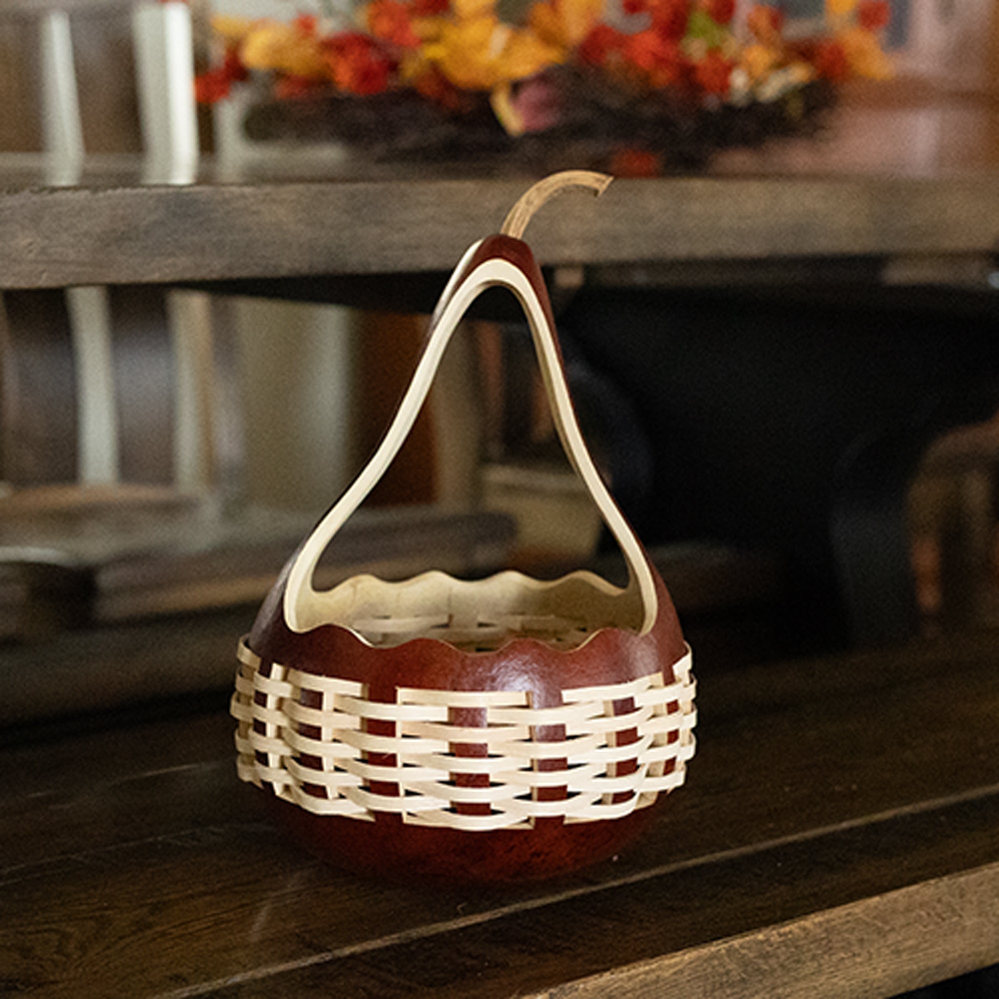 A decorative basket-shaped container made from a brown gourd with a natural woven reed and gourd handle, placed on a dark wooden surface in an indoor setting.