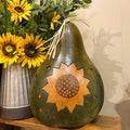A decorative carved gourd sculpture shaped like a pear, featuring a large sunflower design on the front, displayed on a wooden shelf next to a vase of sunflowers.