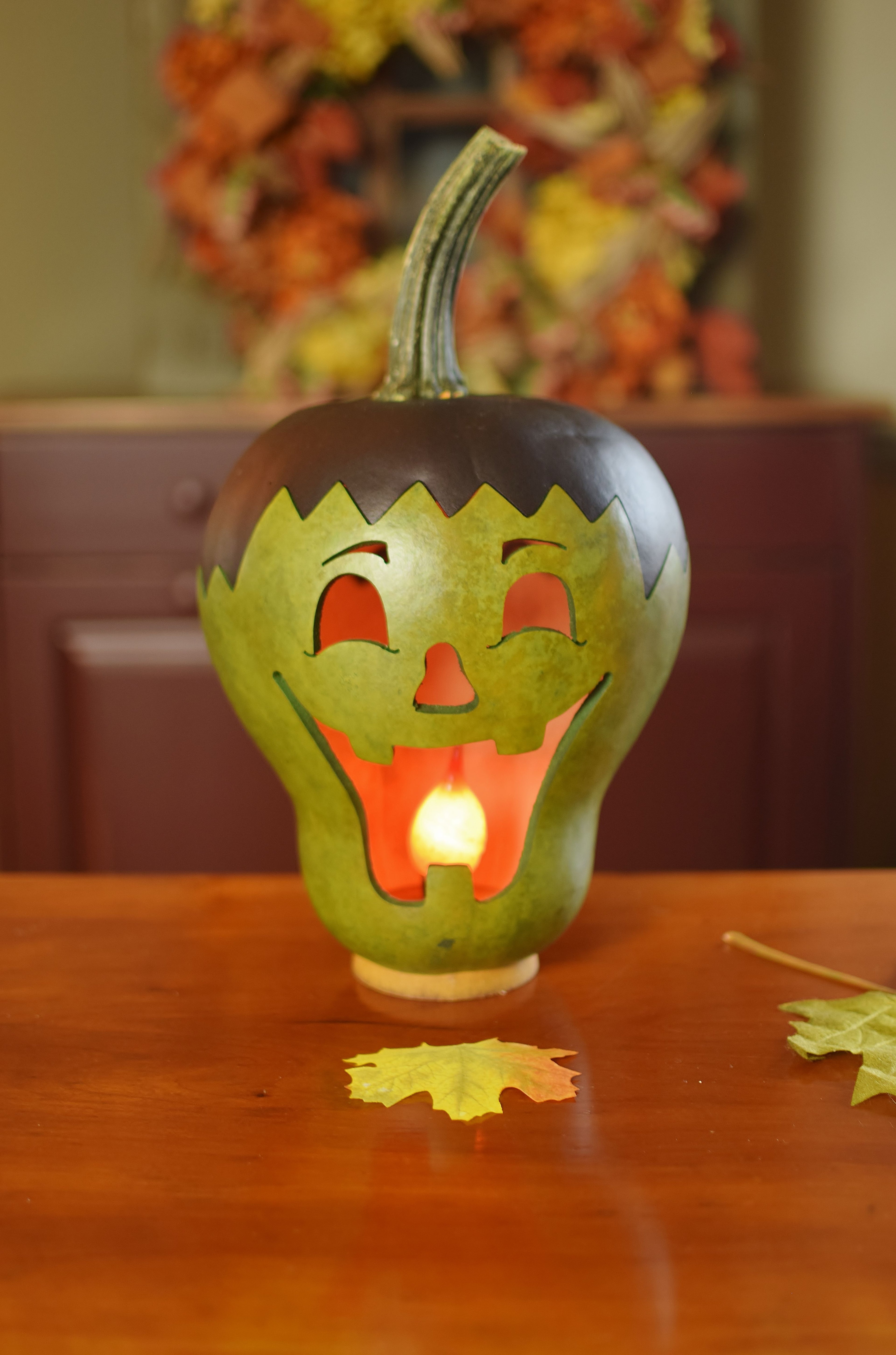 A carved green gourd pumpkin lantern with a glowing tea light inside, placed on a wooden table with a blurred autumn-themed background.