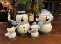 Four handcrafted wooden snowman figurines in different sizes, decorated with felt hats, scarves, and bases, displayed on a wooden table with a pine garland in the background.