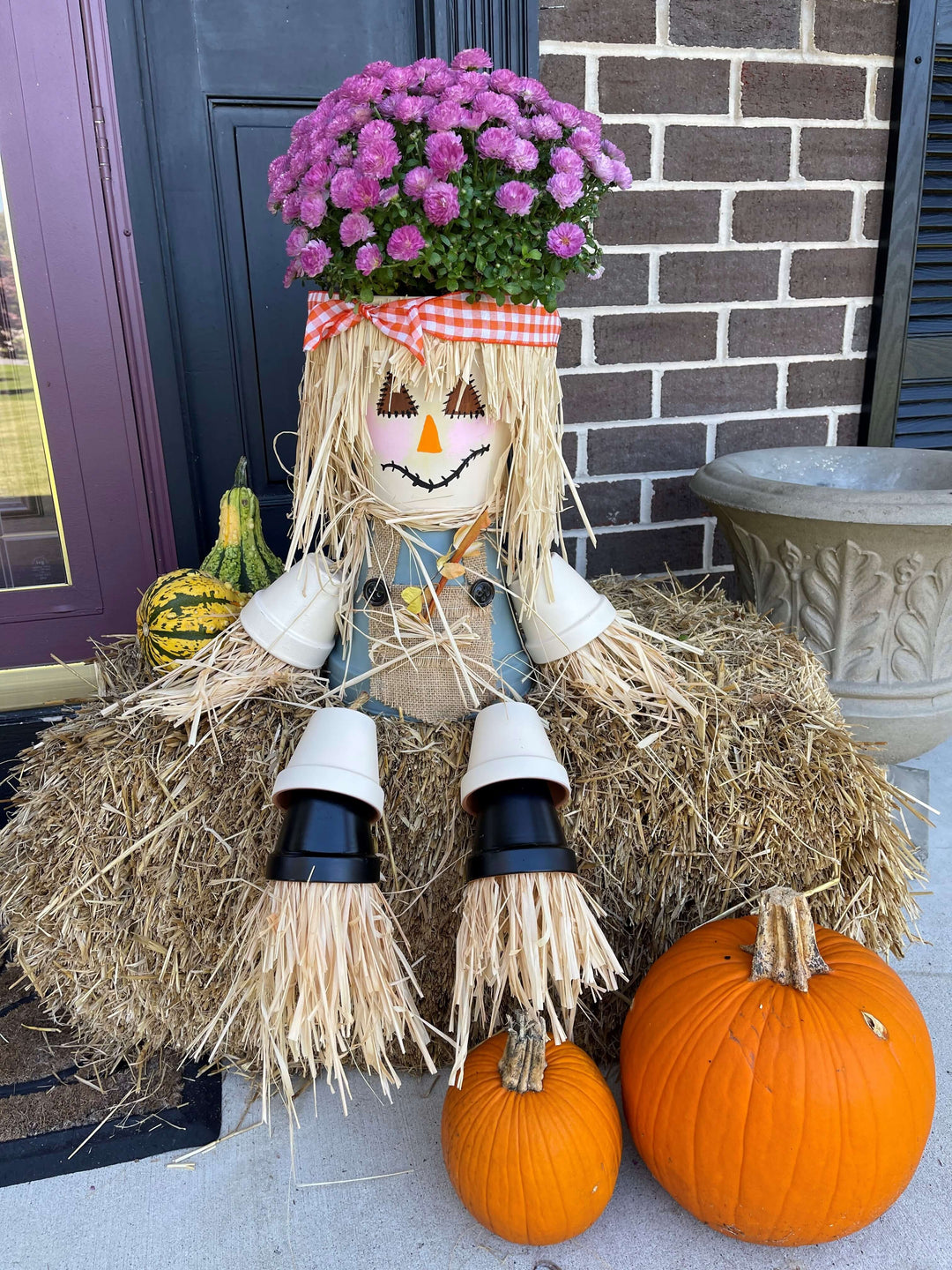 Scarecrow with purple mums on head, sitting on a small hay bale 
